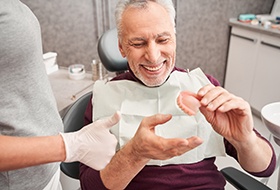 Man in maroon sweater in dental chair smiling holding dentures