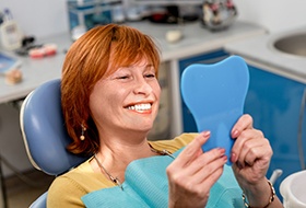 Woman with red hair smiling at her reflection in handheld tooth-shaped mirror